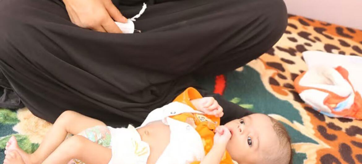 An infant lies on a patterned rug next to an adult in black clothing.