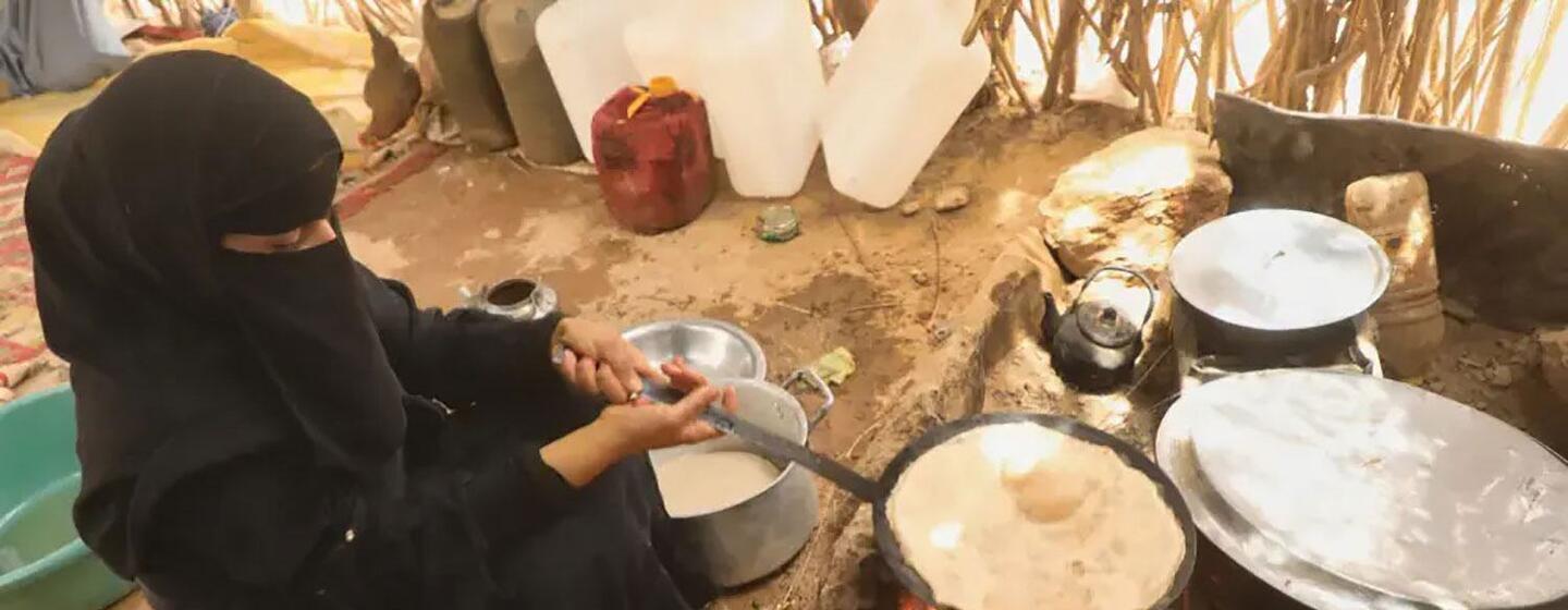 A Yemeni woman in a black niqab kneads dough by an open fire inside a simple rural hut, surrounded by pots and water containers.