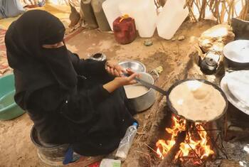 A Yemeni woman in a black niqab kneads dough by an open fire inside a simple rural hut, surrounded by pots and water containers.