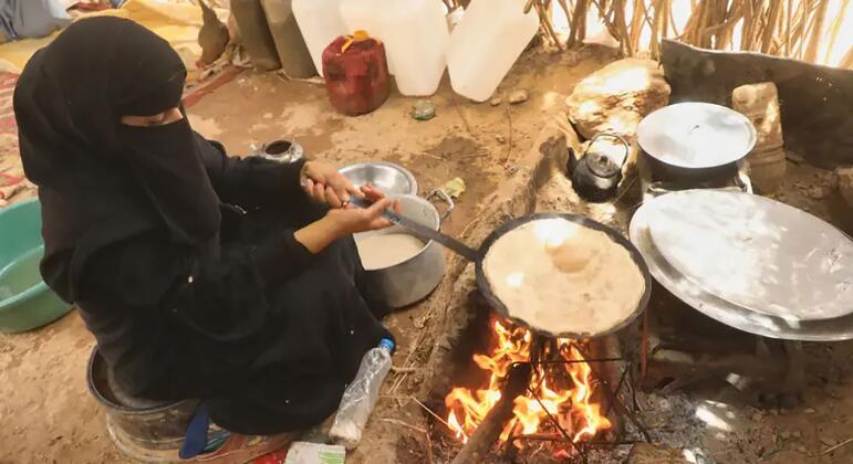 A Yemeni woman in a black niqab kneads dough by an open fire inside a simple rural hut, surrounded by pots and water containers.