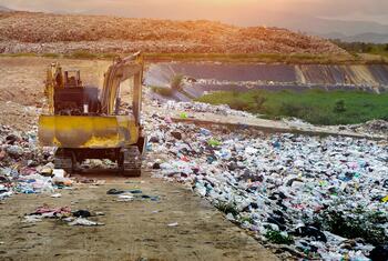 Une pelleteuse jaune travaille au milieu d'amas de déchets plastiques et d'ordures ménagères sur un site de gestion des déchets, avec des collines en arrière-plan.