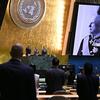 Skrini mbili kubwa zinaonyesha picha nyeusi na nyeupe ya Mfalme Harald wa Tano wa Norway akiwa amevalia sare za sherehe huku wajumbe wakisimama katika ukumbi wa Baraza Kuu UN .