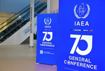 Two tall blue banners displaying the IAEA logo, a large 70 numeral, and GENERAL CONFERENCE text stand in a bright conference hall with escalators and a branded pillar.