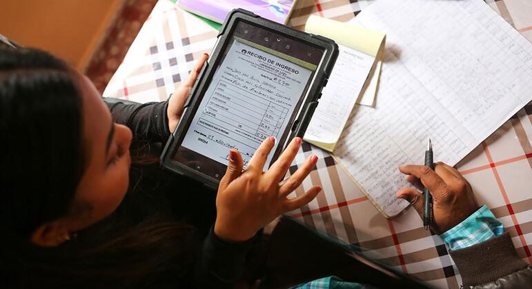 Una mujer latina revisa un recibo de ingresos digital en una tableta mientras un niño escribe en un cuaderno sentado a una mesa.