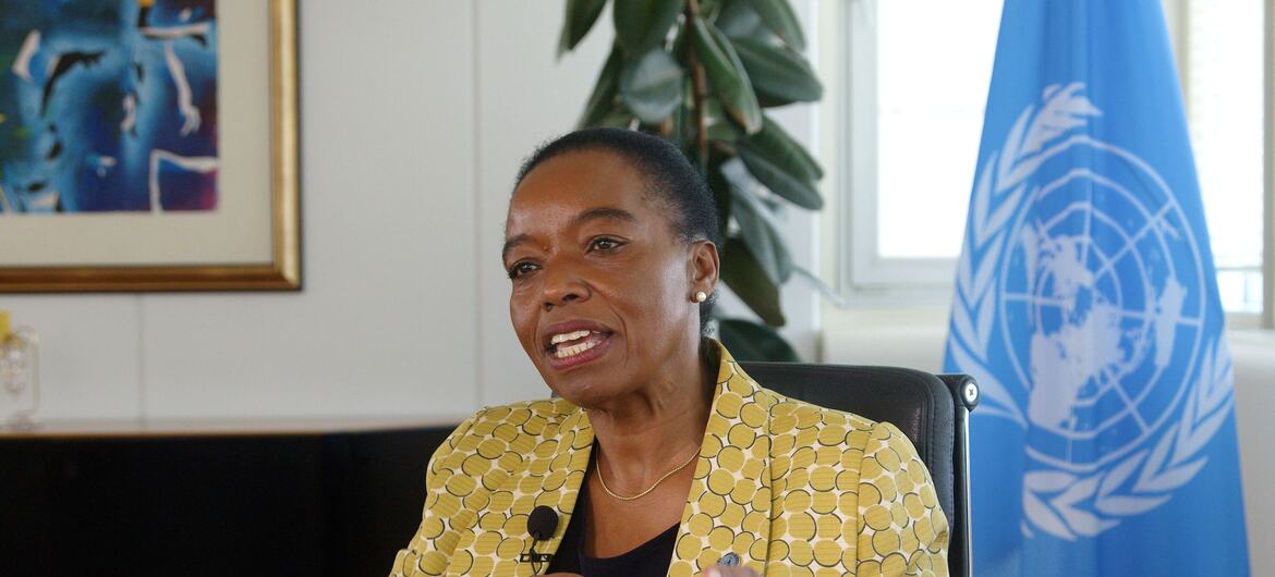 تجلس مونيكا جوما، المديرة التنفيذية UNODC ، على كرسي مرتديةً سترة صفراء منقوشة، وتشير بيدها أثناء حديثها خلال مقابلة، وخلفها علم United Nations .