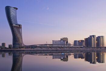 The Abu Dhabi National Exhibition Centre with its curved glass tower and surrounding buildings reflected in calm water at dusk.