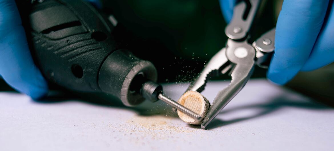 Hands in blue nitrile gloves clamp a small cylindrical sample with locking pliers while a rotary drill bit bores into it, releasing fine dust on a white surface.