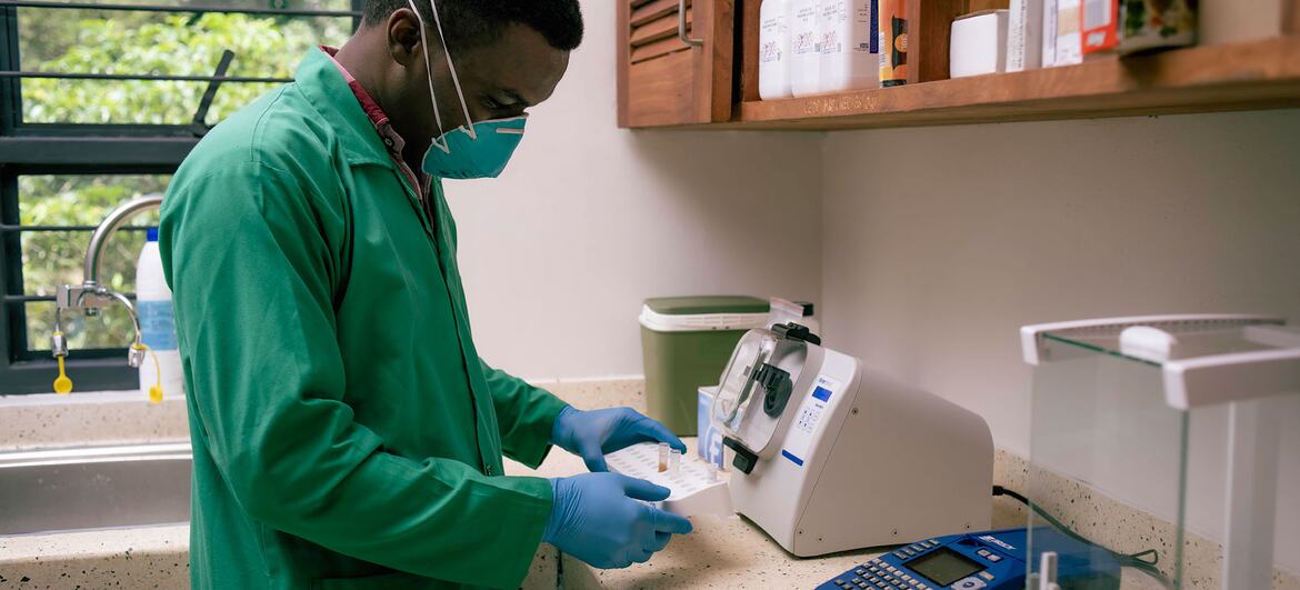 A Black male laboratory technician in a green coat, protective mask and blue gloves handles sample vials at a lab bench equipped with a centrifuge and digital balance.