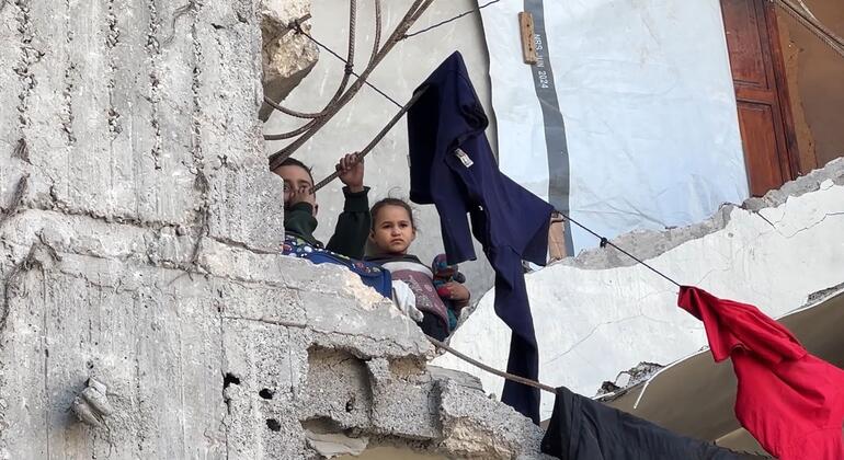 طفلان ينظران من خلف جدار خرساني متضرر في غزة بينما تتدلى الملابس على حبل ممتد عبر الأنقاض.