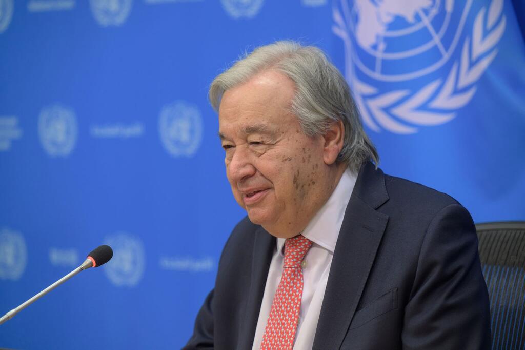 Katibu Mkuu UN António Guterres ameketi mbele ya kipaza sauti wakati wa mkutano na waandishi wa habari, huku mandhari ya bluu UMOJA WA MATAIFA na nembo UN ikionekana nyuma yake.