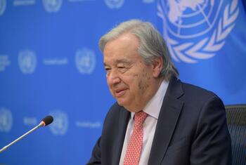 UN Secretary-General António Guterres sits before a microphone during a press briefing, with a blue United Nations backdrop and UN emblem visible behind him.
