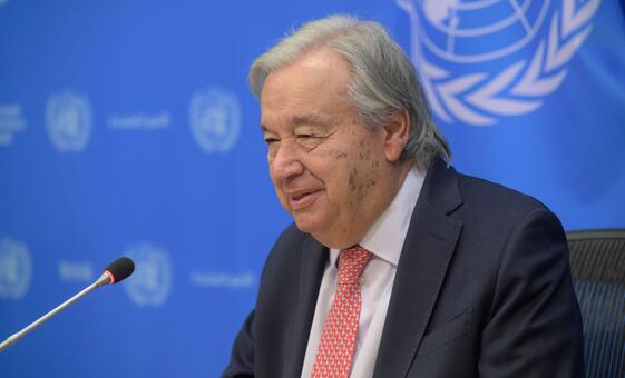 UN Secretary-General António Guterres sits before a microphone during a press briefing, with a blue United Nations backdrop and UN emblem visible behind him.