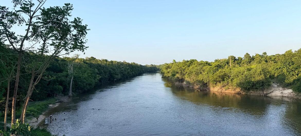 Um rio largo e calmo flui entre margens de densa floresta verde no Brasil, sob um céu azul claro, com pequenas figuras visíveis perto da margem esquerda.