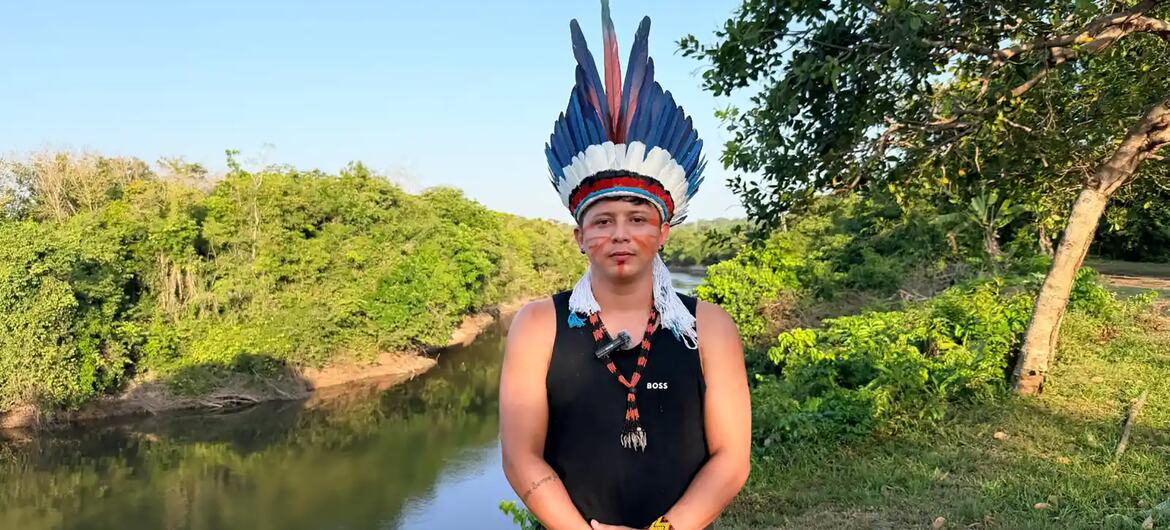 Um jovem usando um cocar de penas, pintura facial e um colar de contas está em pé ao lado de um rio calmo, cercado por árvores verdes.
