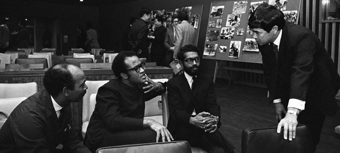 UN decolonization committee members, including FRELIMO representatives Sharfuddine M. Khan and Jorge Rebelo, Tanzania's Salim A. Salim, and Thomas Tanaka, converse in a meeting room with photo displays.