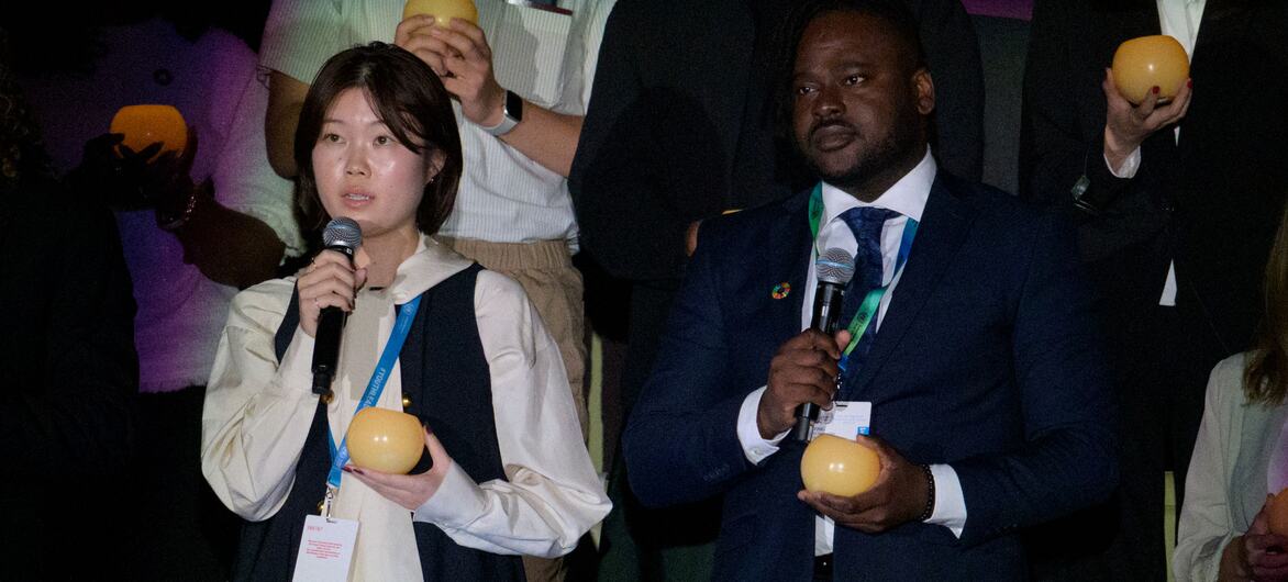 An East Asian woman and a Black man stand at microphones holding yellow fruit during a United Nations event, with other delegates visible behind them.