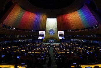 Paneles de luz multicolor cubren las paredes curvas de madera del Salón de la Asamblea General de la ONU , mientras los delegados se sientan en escritorios frente a un podio y dos pantallas que muestran una transmisión de un Momento de los ODS.