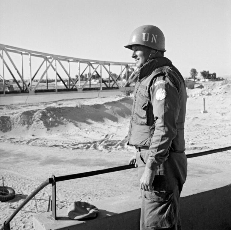 Le capitaine Josep Muller, de l'armée autrichienne, coiffé d'un casque et vêtu d'un uniforme des Nations Unies, se tient à un poste d'observation près du canal de Suez, avec un pont en acier en arrière-plan.
