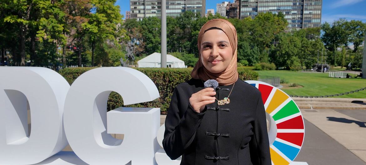 امرأة ترتدي حجاباً بيج اللون ومعطفاً أسود تتحدث في ميكروفون محمول باليد بجانب حروف كبيرة بيضاء لأهداف التنمية المستدامة وعجلة ملونة لأهداف United Nations للتنمية المستدامة.