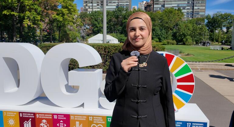 امرأة ترتدي حجاباً بيج اللون ومعطفاً أسود تتحدث في ميكروفون محمول باليد بجانب حروف كبيرة بيضاء لأهداف التنمية المستدامة وعجلة ملونة لأهداف United Nations للتنمية المستدامة.