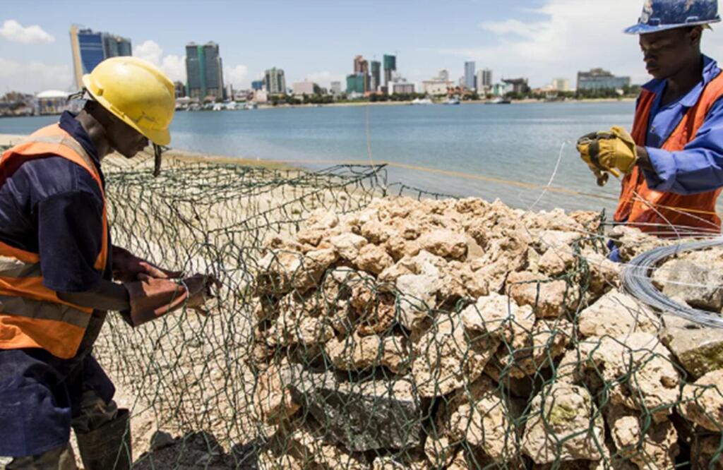 Deux ouvriers du bâtiment noirs, casqués et vêtus de gilets haute visibilité, assemblent des gabions sur une plage de Pangani, avec le port et la silhouette de la ville en arrière-plan.