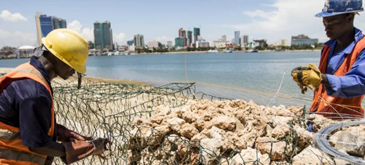 Two Black male construction workers in helmets and high-visibility vests assemble gabion rock cages on a beach in Pangani, with a harbor and city skyline behind.