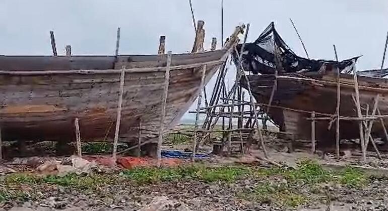 Weathered wooden fishing vessels rest on a sandy shore supported by wooden poles, with dark nets draped over one hull under an overcast sky.