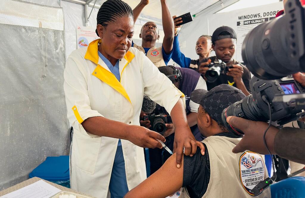 Mfanyakazi wa afya mwanamke mweusi aliyevaa koti jeupe akimdunga sindano mtu aliyevaa fulana ya beige ya kukabiliana na dharura, akiwa amezungukwa na wapiga picha na wafanyakazi wa vyombo vya habari ndani ya hema jeupe.