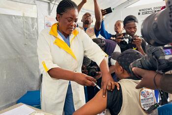 A Black female healthcare worker in a white coat administers an injection to a person wearing a beige emergency response vest, surrounded by photographers and media personnel inside a white tent