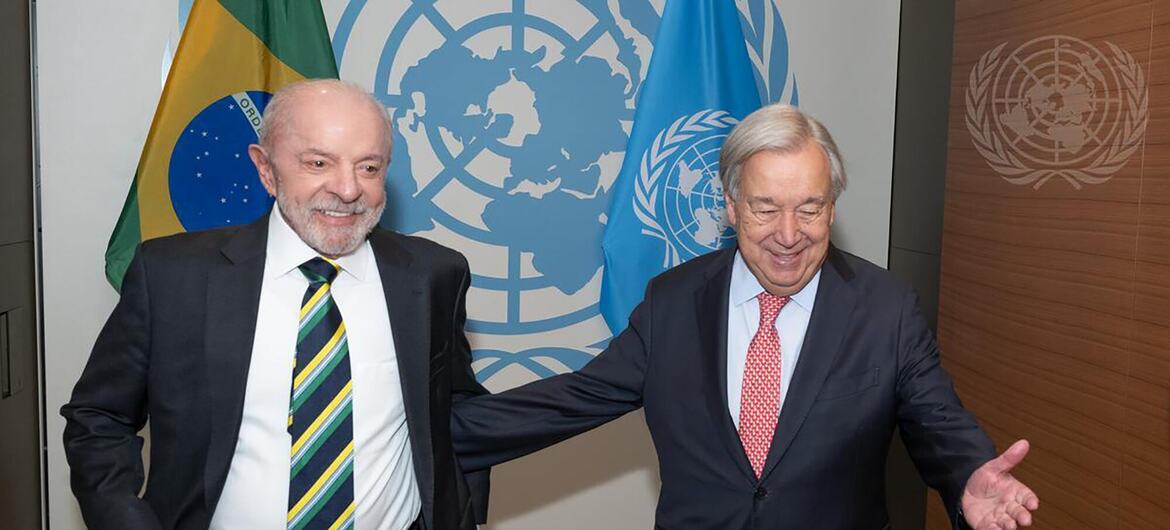 O presidente brasileiro Luiz Inácio Lula da Silva e o secretário-geral da ONU , António Guterres, sorriem diante das bandeiras do Brasil e das Nações Unidas .
