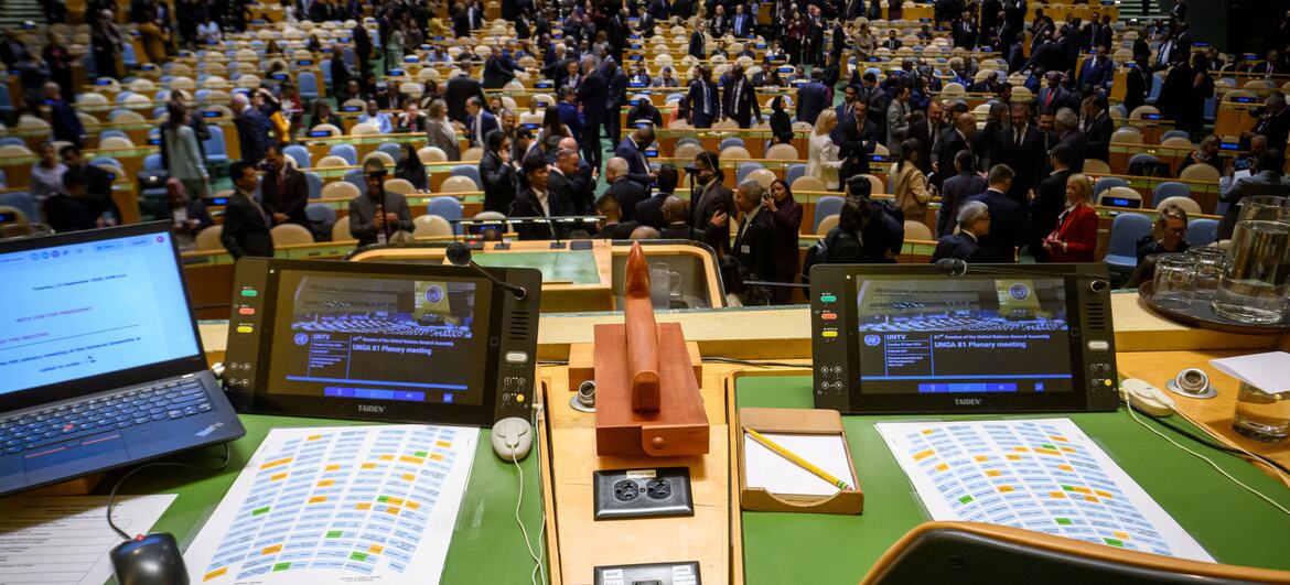 Delegates stand and converse in the United Nations General Assembly Hall while a podium desk holds laptops, monitors, a microphone, and seating charts.