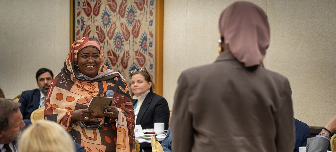 امرأة سوداء مبتسمة ترتدي حجاباً منقوشاً تحمل أوراقاً أثناء حديثها في اجتماع رسمي يجلس فيه الحضور؛ ويجلس الحاضرون الآخرون، بمن فيهم امرأة ترتدي بدلة بنية اللون وحجاباً وردياً وظهرها للكاميرا، حول طاولة.