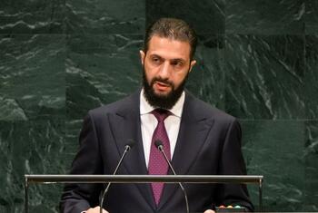 أحمد الشرع، رئيس الجمهورية العربية السورية، يلقي كلمة من على منصة United Nations أمام ميكروفونات وشعار الأمم المتحدة على خلفية من الرخام الأخضر.