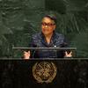 A Black woman in glasses and a patterned blue jacket speaks at a United Nations podium bearing the UN emblem, with green marble walls behind her during a General Assembly session.