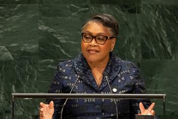 A Black woman in glasses and a patterned blue jacket speaks at a United Nations podium bearing the UN emblem, with green marble walls behind her during a General Assembly session.