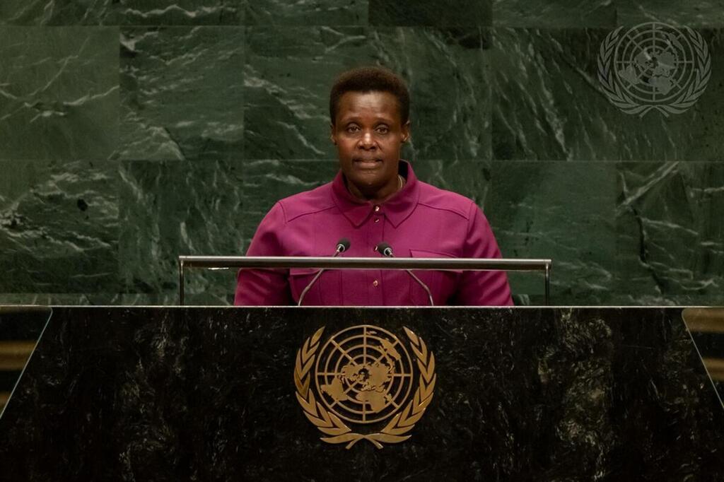 Mwanamke mweusi aliyevaa kanzu ya magenta amesimama kwenye jukwaa la marumaru nyeusi lenye nembo UMOJA WA MATAIFA , akizungumza kwenye maikrofoni mbili dhidi ya ukuta wa marumaru ya kijani yenye nembo UN .