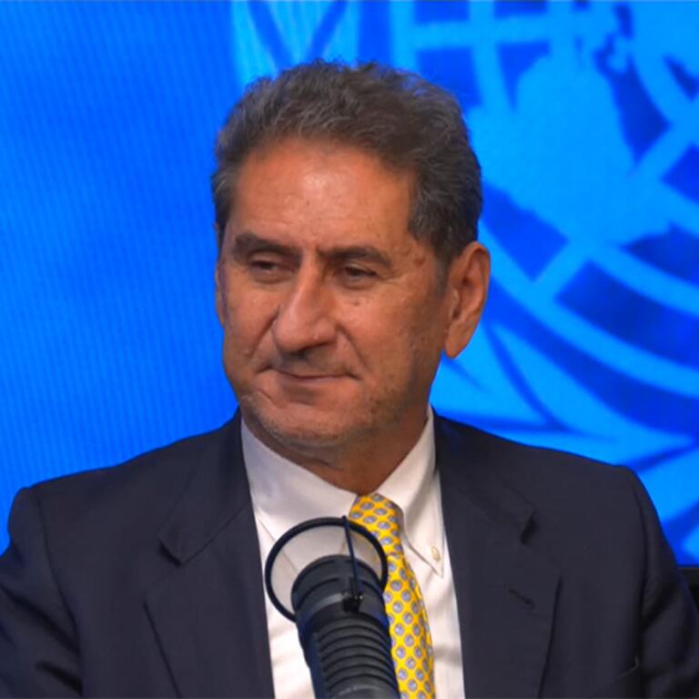 Un hombre con traje oscuro y corbata amarilla habla por un micrófono en un evento de las Naciones Unidas , con el emblema de la ONU proyectado en una pantalla azul detrás de él.