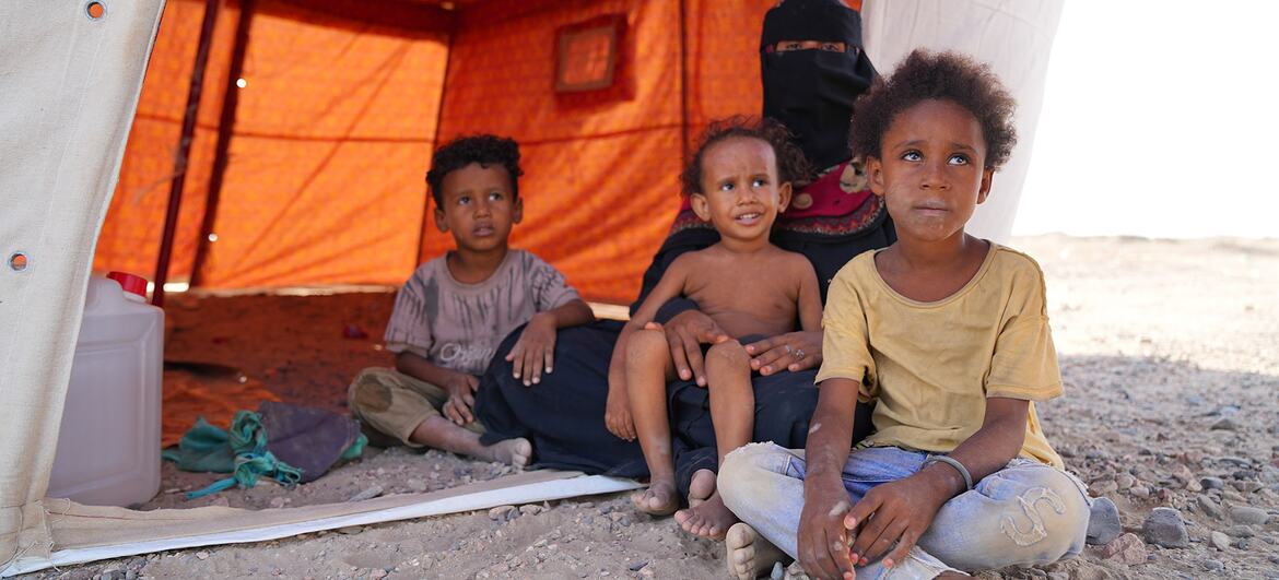 A Yemeni mother wearing a niqab sits with four young children at the entrance of an orange emergency shelter tent in Al-Firdous IDP camp, Aden, Yemen.