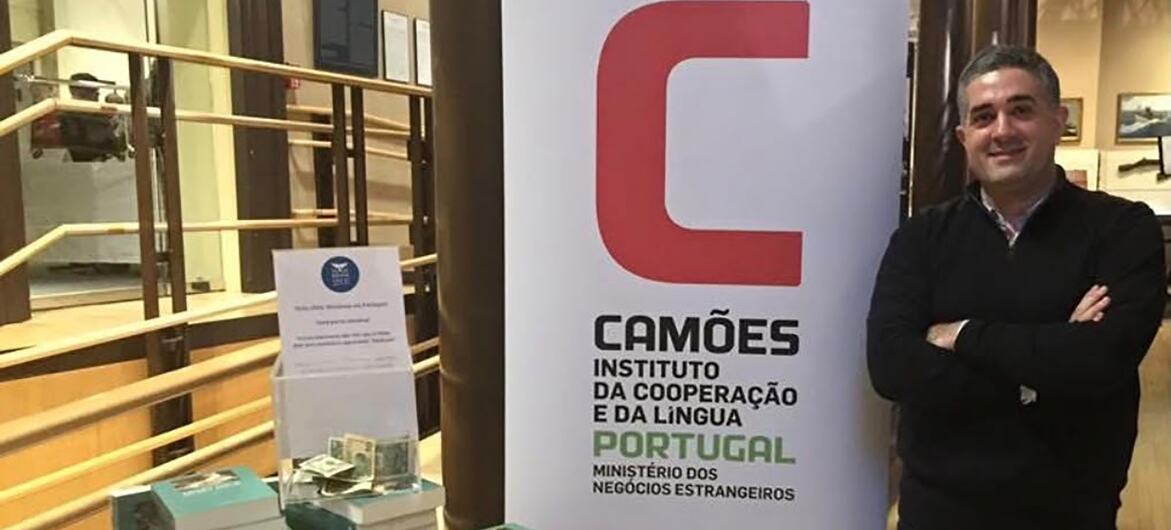 João Caixinha, um português de meia-idade, de cabelos grisalhos e óculos, aparece em frente a um fundo branco com o logotipo do Instituto Camões e um texto parcial relacionado à língua portuguesa e a Lisboa.