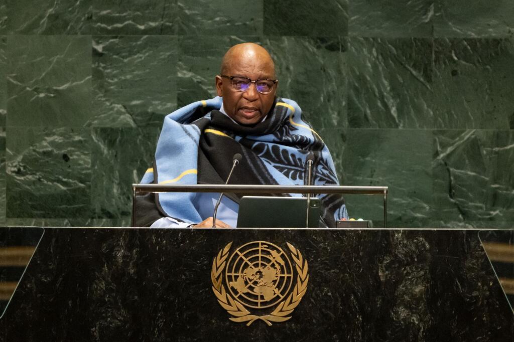 Mwanamume Mweusi aliyevaa miwani na blanketi yenye muundo wa bluu na nyeusi akizungumza kwenye jukwaa la UMOJA WA MATAIFA akiwa na nembo ya dhahabu UN , maikrofoni, na kibao mbele yake.