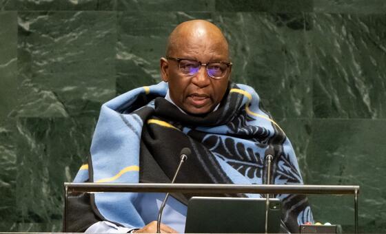 Mwanamume Mweusi aliyevaa miwani na blanketi yenye muundo wa bluu na nyeusi akizungumza kwenye jukwaa la UMOJA WA MATAIFA akiwa na nembo ya dhahabu UN , maikrofoni, na kibao mbele yake.