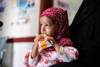A young Yemeni girl in a red floral headscarf eats from an orange nutrition packet while seated beside an adult in a black abaya at a clinic.