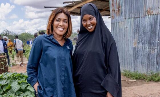 Wanawake wawili wanasimama karibu na kutabasamu nje; mmoja amevaa shati la bluu nyeusi lenye nembo ndogo, huku mwingine amevaa kifuniko cheusi cha kichwa. Mimea yenye majani, njia ya vumbi, watu wa mbali, na ukuta wa chuma uliochakaa unaonekana nyuma yao.
