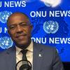 Um homem negro, de terno cinza e óculos, fala ao microfone diante de um fundo azul com a inscrição "ONU NEWS" e emblemas repetidos das Nações Unidas .