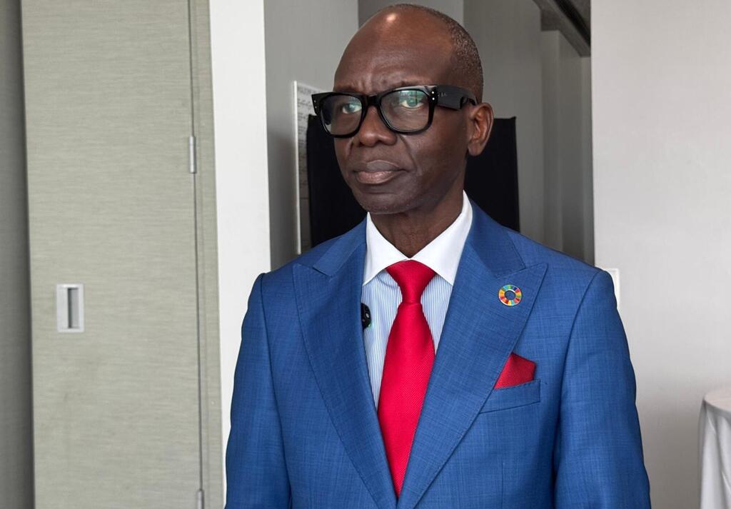 Profesa Janabi, mwanaume Mweusi aliyevaa miwani yenye fremu nyeusi, suti ya bluu, shati jeupe na tai nyekundu, amesimama kwenye korido ya ndani akiwa amevaa pini ya mviringo yenye rangi mbalimbali.