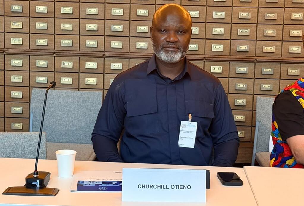 Churchill Otieno ameketi kwenye meza ya mikutano akiwa na bamba la jina, kipaza sauti, na hati UN wakati wa mahojiano. Amevaa shati jeusi la rangi ya samawati, na safu za droo za kadi nyuma yake.
