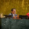 A man in a grey suit and glasses speaks at a green marble desk in the UN General Assembly hall, while a woman in a red top works on a laptop beside him.