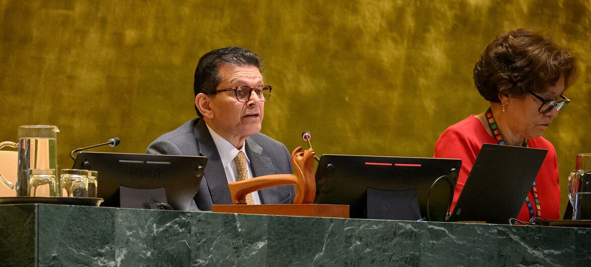 A man in a grey suit and glasses speaks at a green marble desk in the UN General Assembly hall, while a woman in a red top works on a laptop beside him.