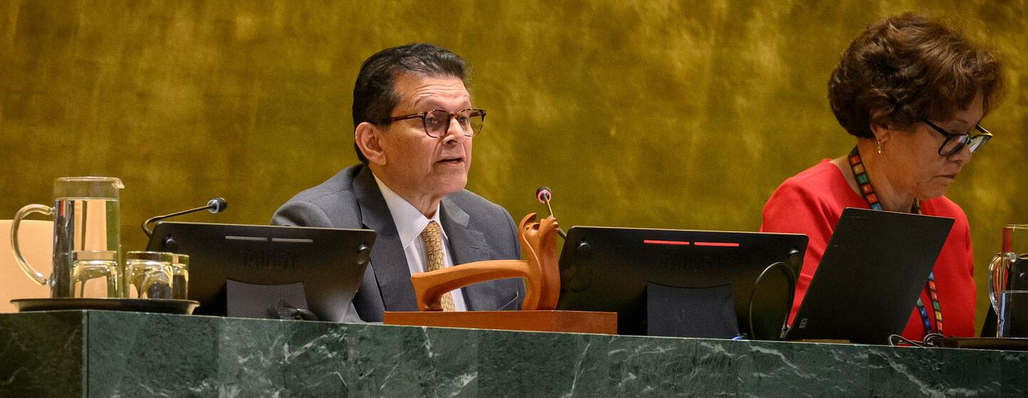 A man in a grey suit and glasses speaks at a green marble desk in the UN General Assembly hall, while a woman in a red top works on a laptop beside him.