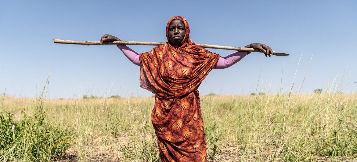 Une femme noire soudanaise se tient dans un champ aride près de Guriesha, dans l'État de Gedaref, tenant une longue houe en bois sur ses épaules, vêtue d'une robe à motifs orange et d'un foulard sur la tête.